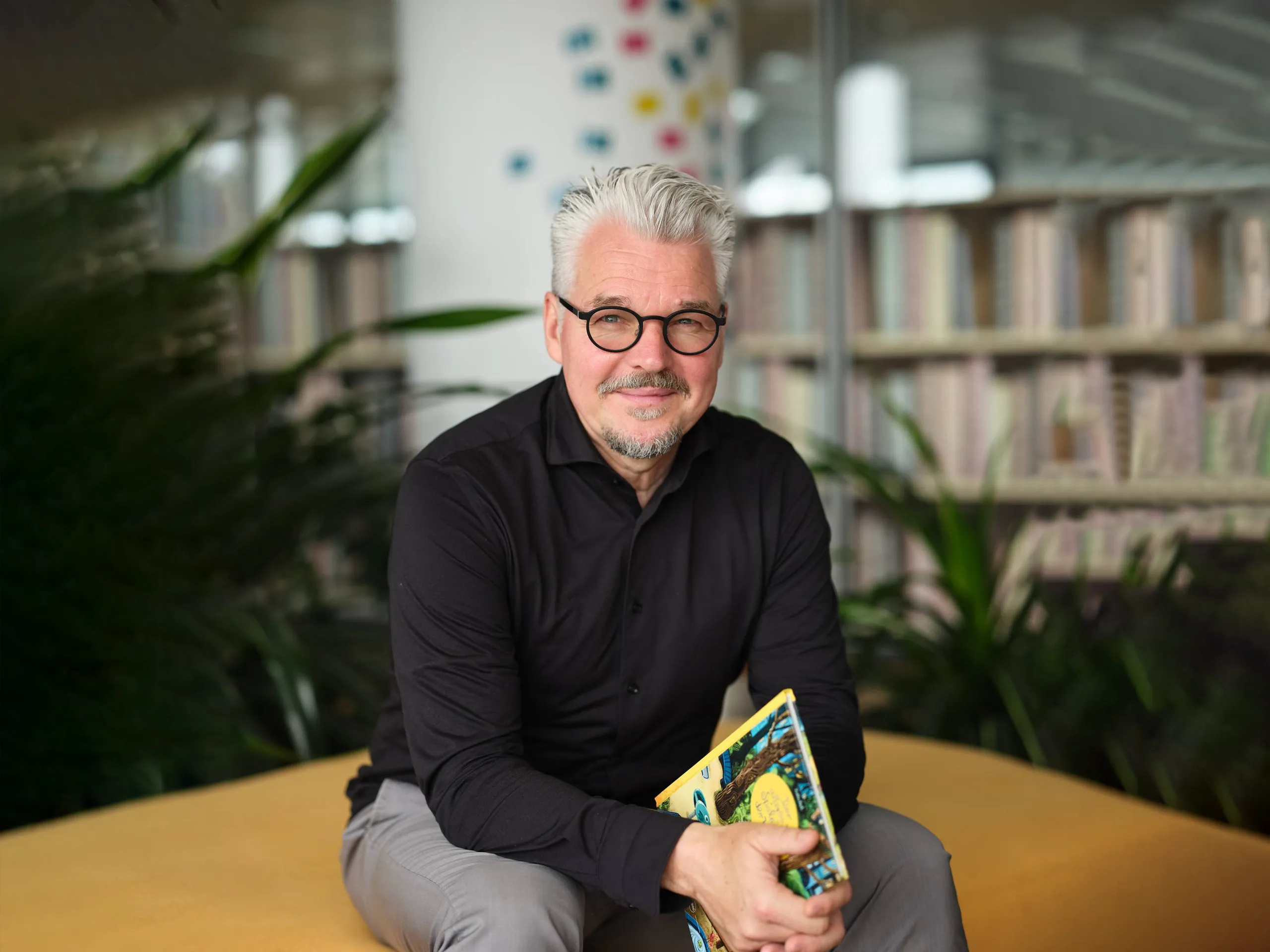 Person sitzt auf einem gelben Sitzmöbel in einem modernen Raum mit Bücherregalen im Hintergrund. Die Person trägt ein schwarzes Hemd und hält ein bunt illustriertes Kinderbuch mit dem Titel „Der allerkleinste Spielplatz der Welt!“ in den Händen. Im Hintergrund sind Pflanzen und eine Glaswand mit bunten Punkten sichtbar.