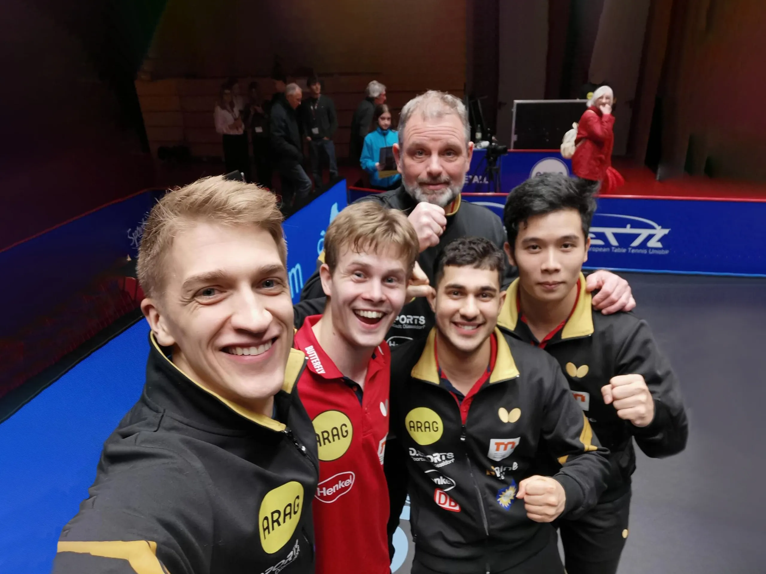 Gruppe von Tischtennisspielern steht eng zusammen in einer Sporthalle und posiert für ein Foto. Die Spieler tragen schwarze und rote Trikots mit Sponsorenlogos, darunter „ARAG“ und „Butterfly“. Im Hintergrund sind Tischtennisplatten, blaue Banden und einige Personen zu sehen.