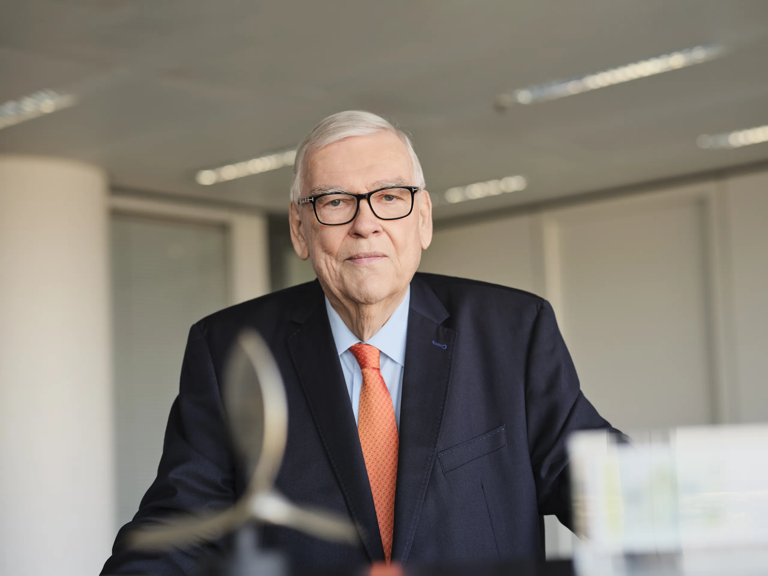 Person in einem dunklen Anzug mit orangefarbener Krawatte steht in einem modernen Büro. Im Vordergrund ist eine unscharfe Skulptur zu sehen, im Hintergrund helle Wände und Deckenbeleuchtung.