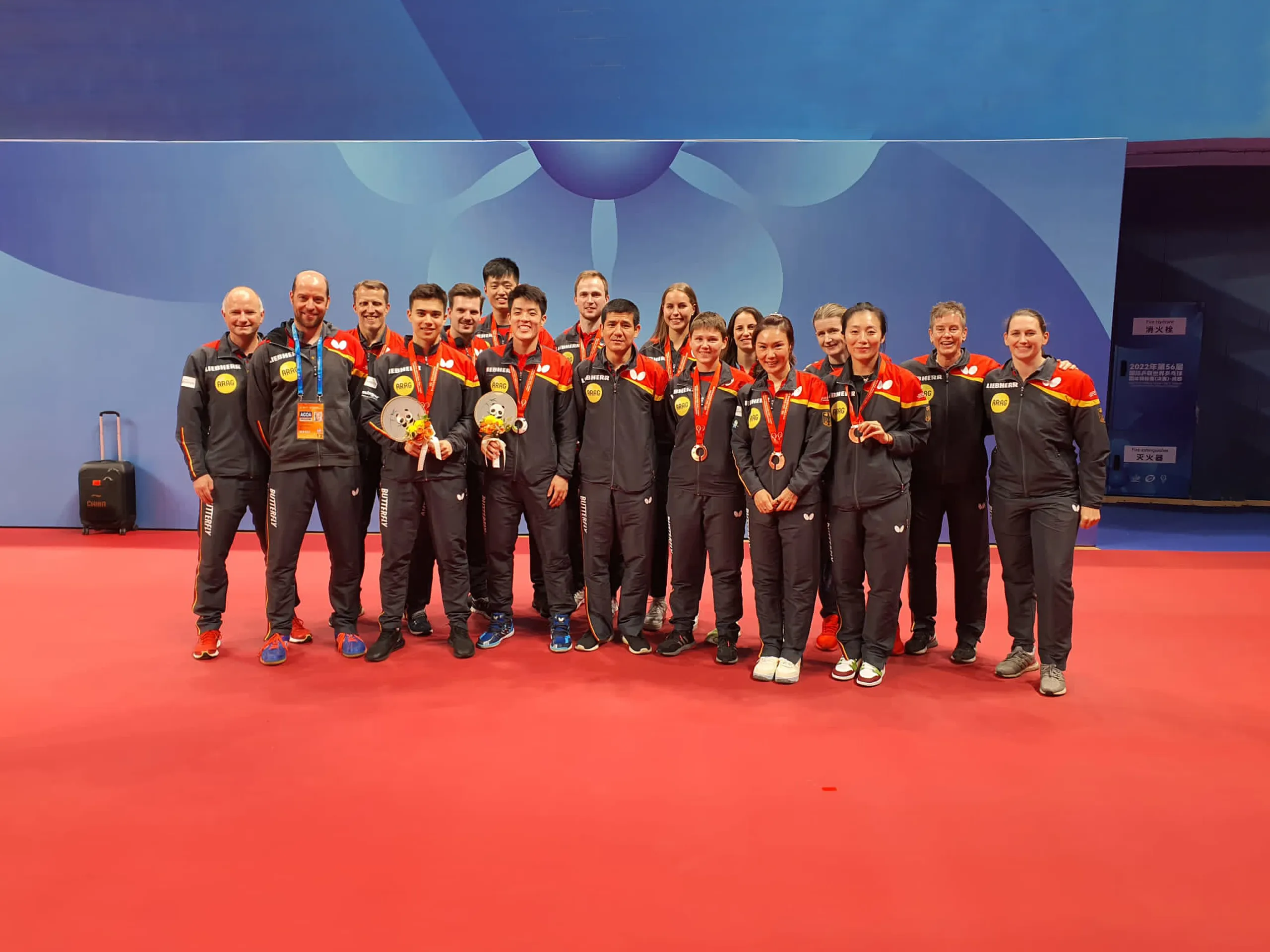 Eine Gruppe von Tischtennisspielern steht auf rotem Boden vor einer blauen Wand und posiert für ein Teamfoto. Alle tragen dunkelgraue Trainingsanzüge mit roten und gelben Akzenten sowie Sponsorenlogos, darunter „ARAG“. Mehrere Personen halten Medaillen und Pokale in den Händen. Im Hintergrund ist ein Koffer sichtbar.
