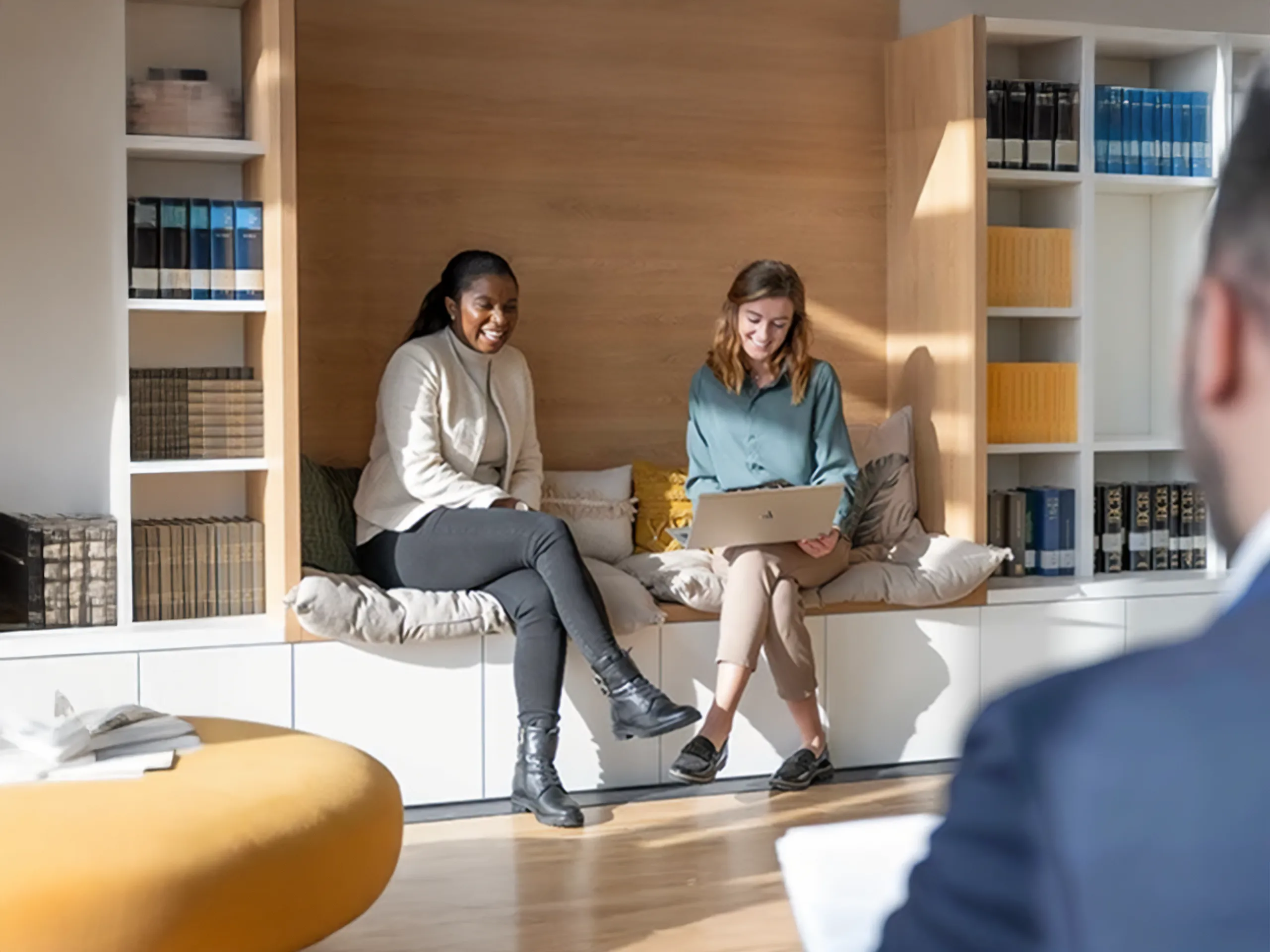 Zwei Frauen sitzen lächelnd in einer modernen Büroecke und schauen auf einen Laptop.