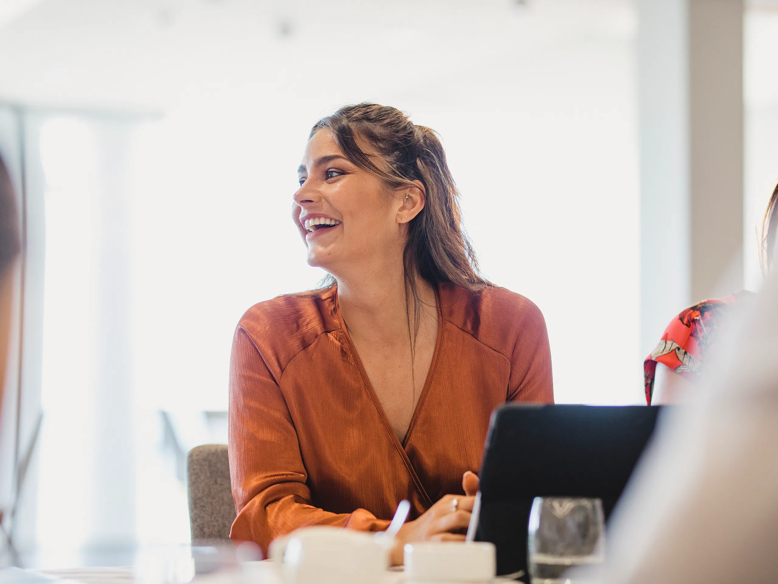 Lachende Frau sitzt im Meeting und arbeitet an einem Laptop.