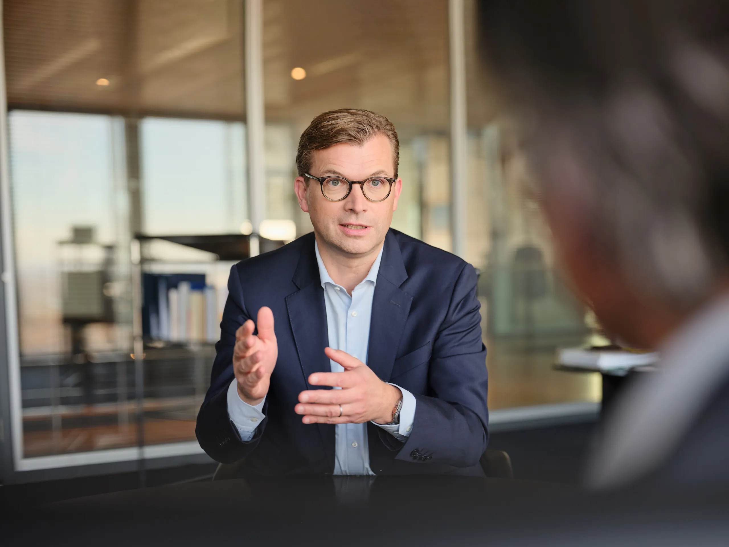 Person in formeller Kleidung sitzt in einem modernen Büro und gestikuliert während eines Gesprächs. Im Hintergrund sind Glaswände und Regale mit Ordnern sichtbar, die eine professionelle Arbeitsumgebung zeigen.
