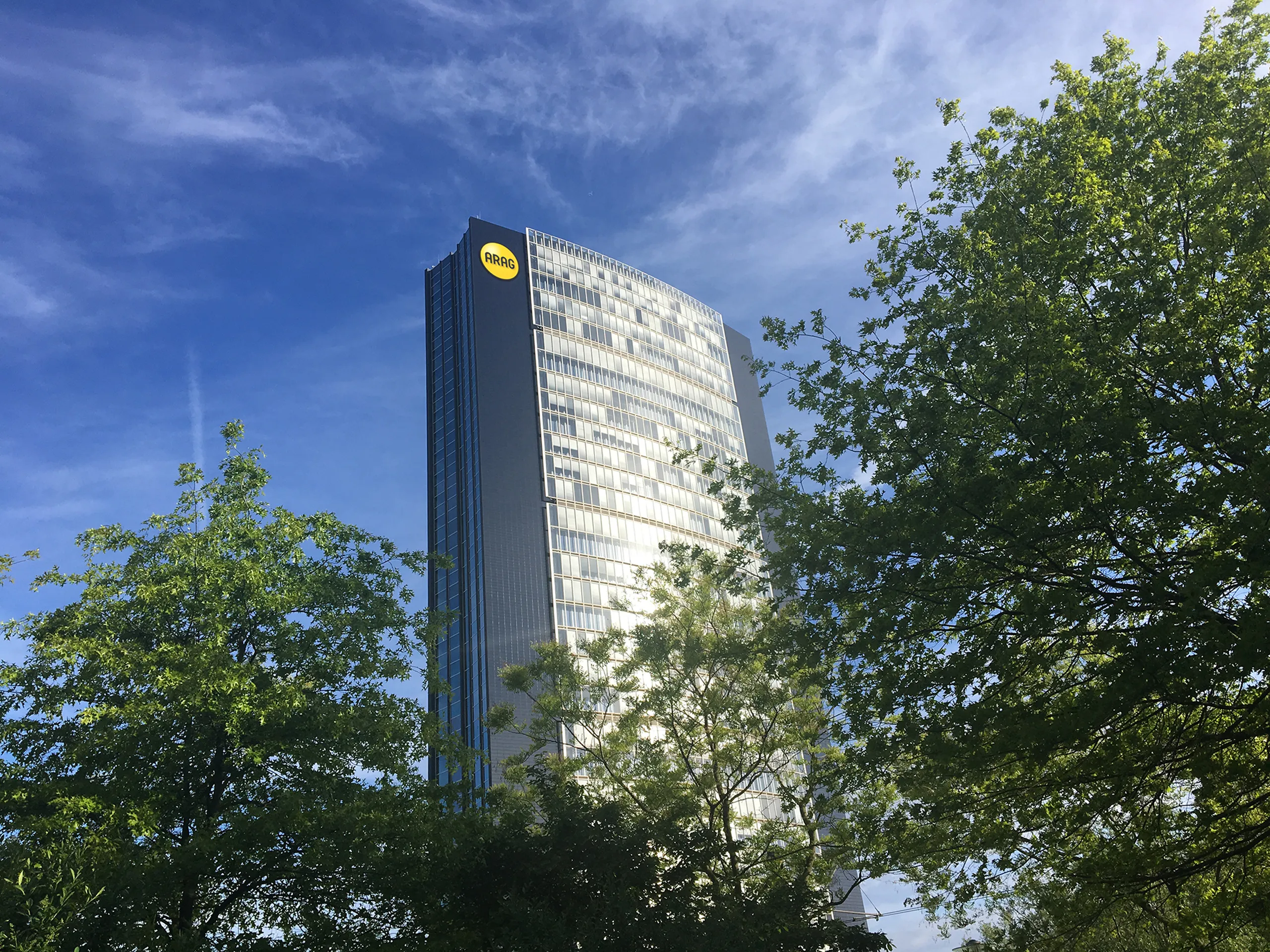 Hohes, modernes Bürogebäude mit Glasfassade und dem gelben ARAG-Logo oben an der Fassade. Im Vordergrund sind grüne Bäume zu sehen, darüber ein blauer Himmel mit leichten Wolken.