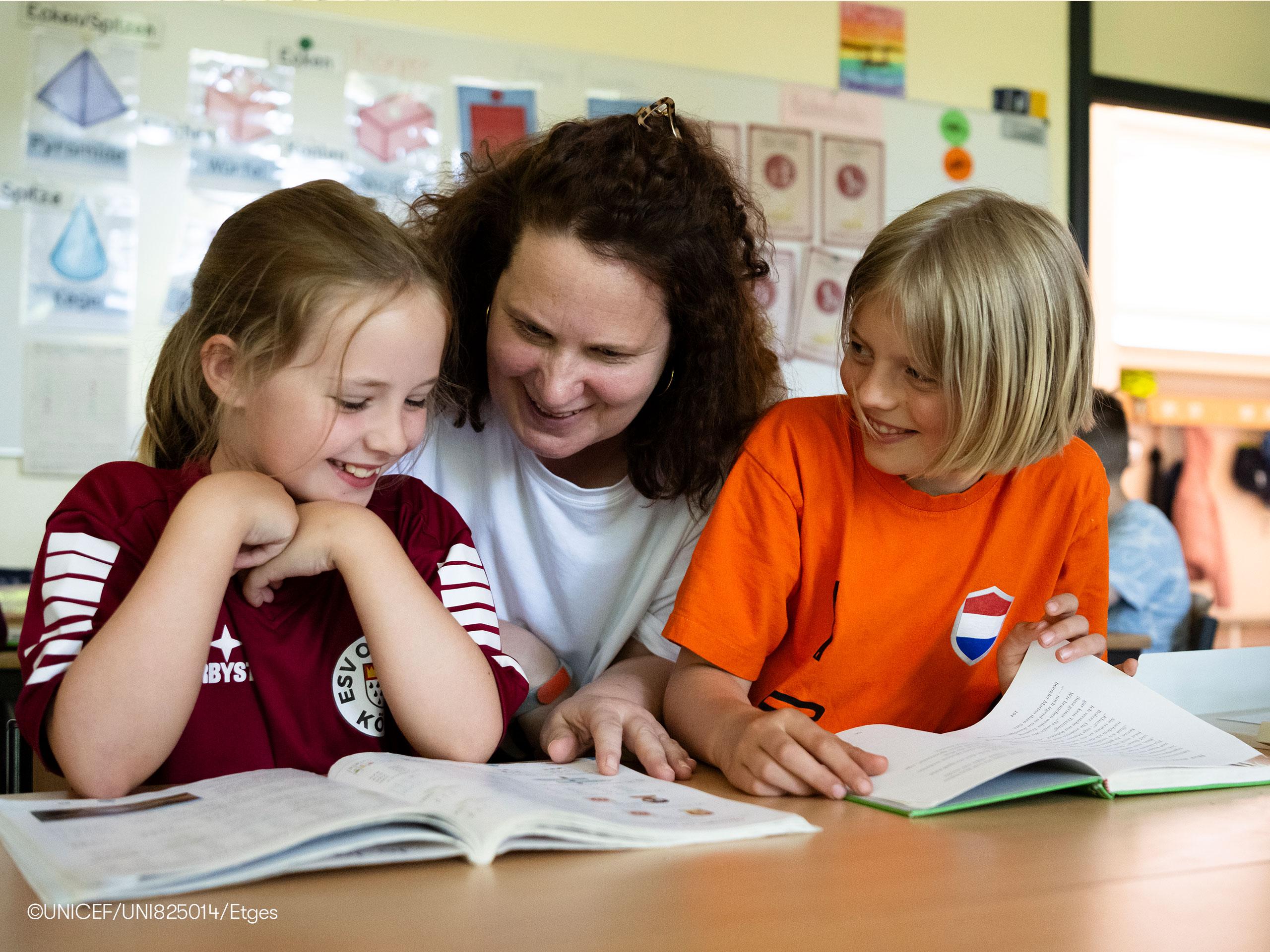 Eine erwachsene Person sitzt an einem Tisch mit zwei Kindern und betrachtet gemeinsam mit ihnen aufgeschlagene Schulbücher in einem Klassenraum. Im Hintergrund sind Lernmaterialien an der Wand zu sehen.