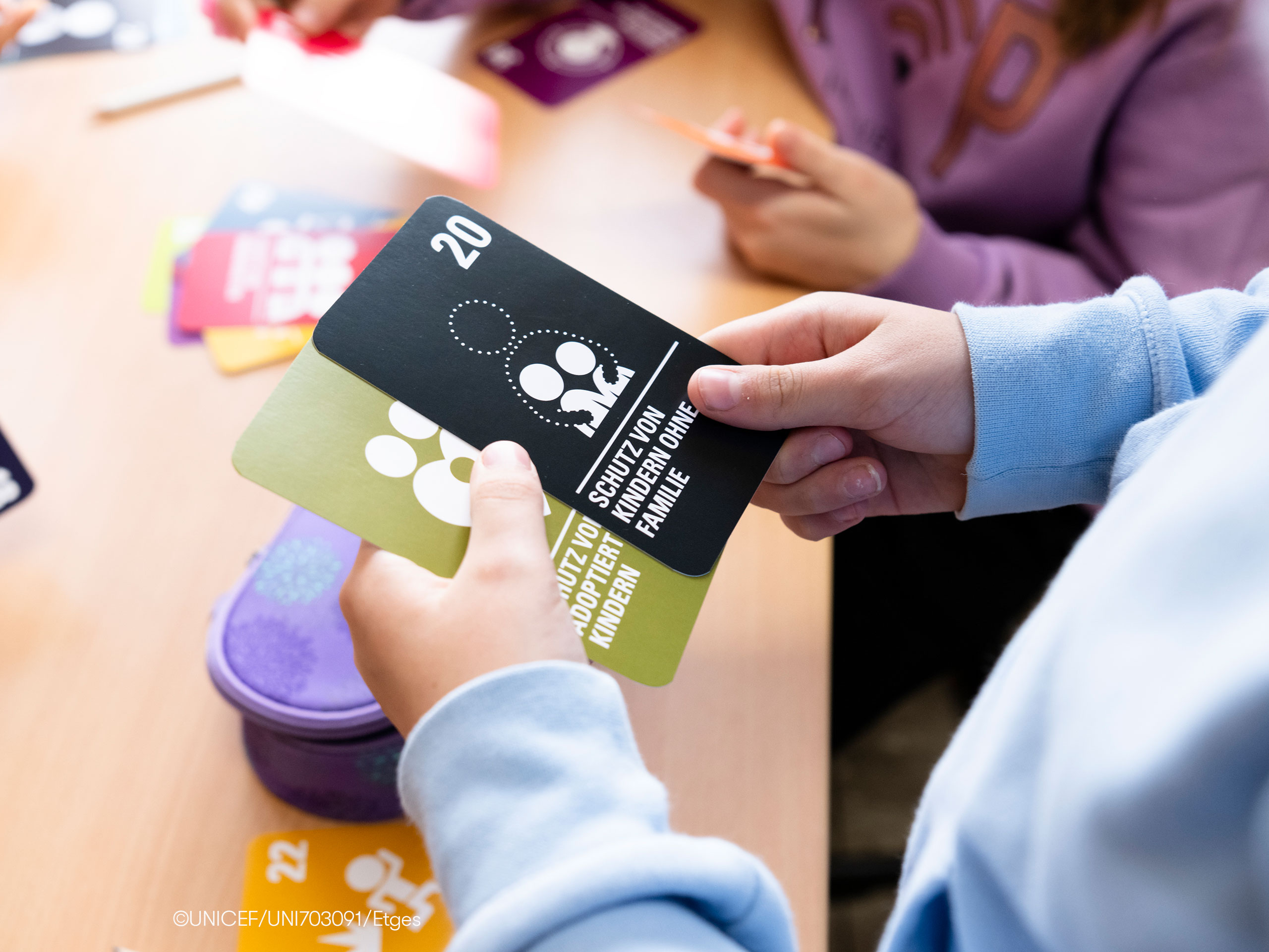 Ein Kind hält mehrere Karten mit Zahlen und Symbolen in den Händen, während im Hintergrund weitere Kinder an einem Tisch sitzen und mit Materialien arbeiten.