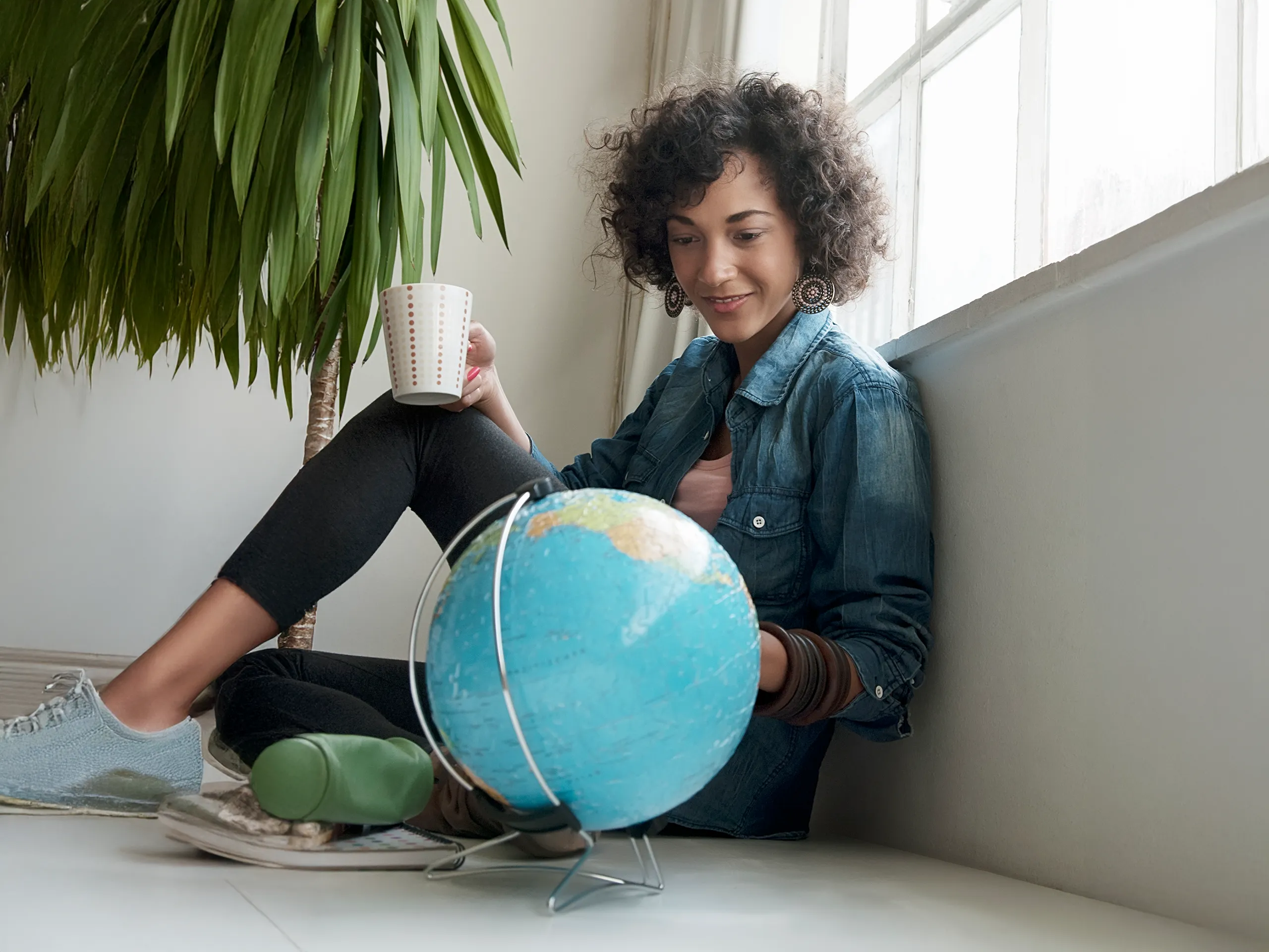 Frau sitzt am Fenster, hält eine Tasse und betrachtet einen Globus.